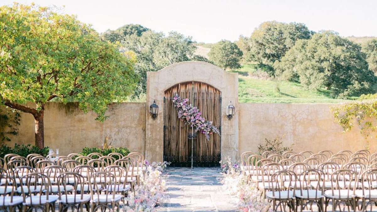 Outdoor wedding venue with mission-style adobe walls and large rustic wood gate covered with flowers at The Club at Pasadera Monterey